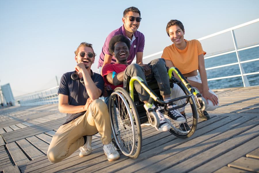 Un groupe d'amis, dont une personne en fauteuil roulant, riant ensemble en vacances au bord de l'eau